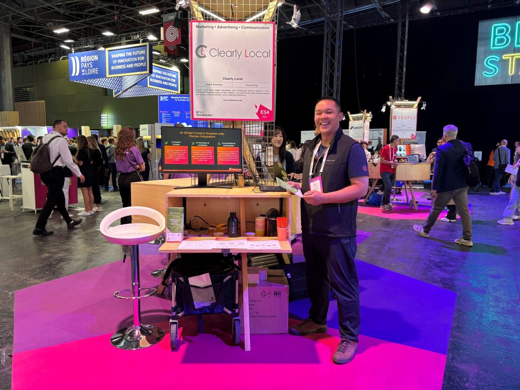 A smiling man stands in front of a Clearly Local exhibition booth at a tech event, surrounded by people and startup displays. The booth features a screen, promotional materials, and a bright pink and purple floor, with signs and lights from other exhibitors in the background.