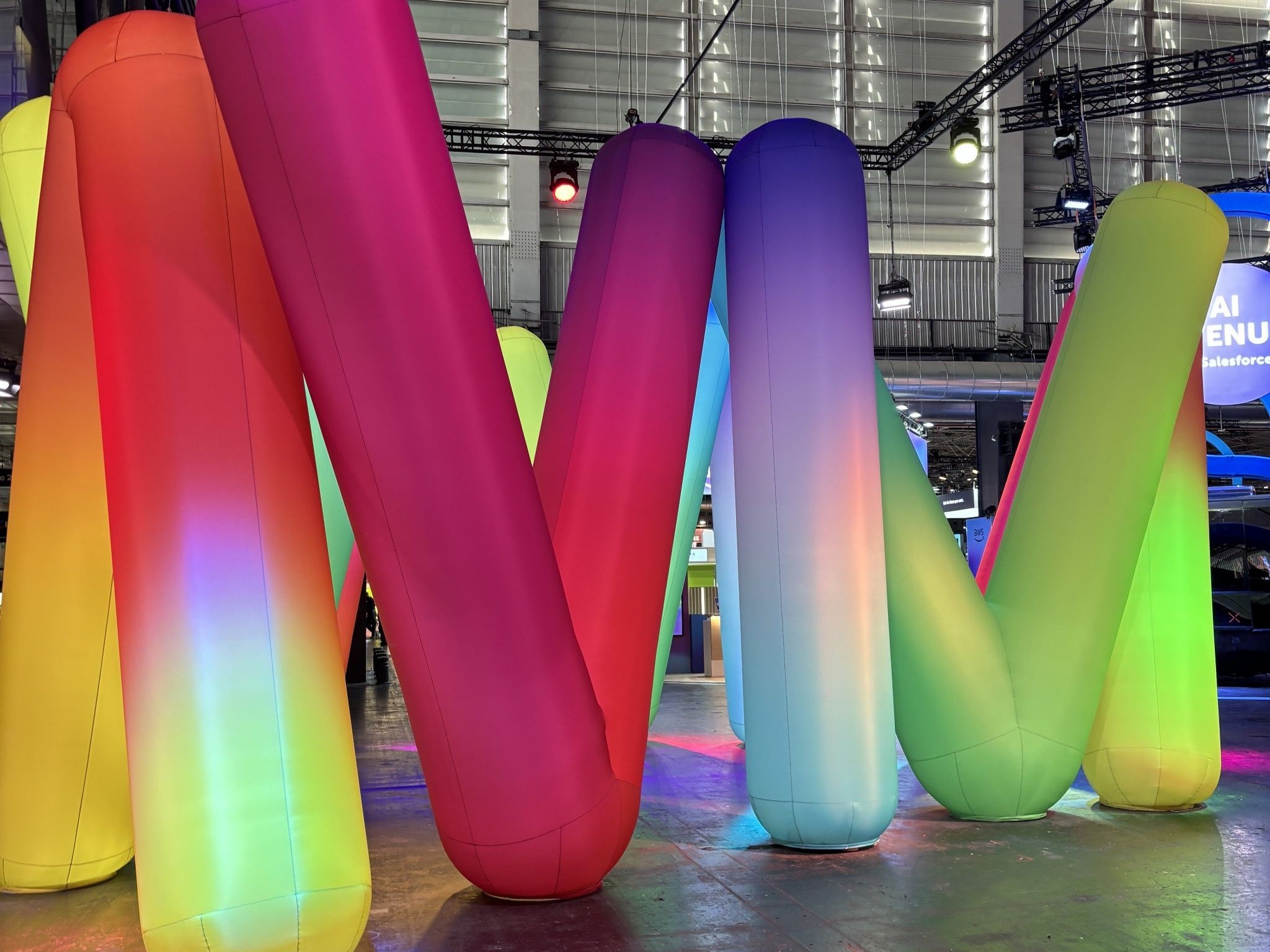 Large, colorful inflatable tubes in gradient hues of red, orange, pink, purple, blue, green, and yellow form a playful installation at an indoor event space with industrial-style lighting and high ceilings.