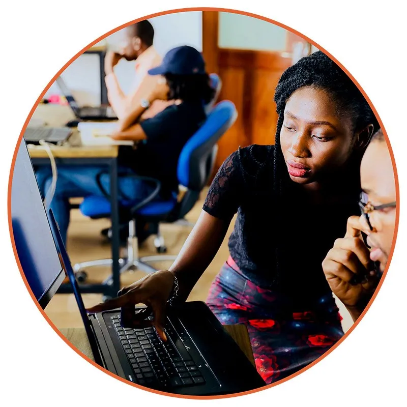 A woman is focused, pointing at a laptop screen, guiding a colleague in a vibrant office. Two others work in the background, creating a collaborative atmosphere.