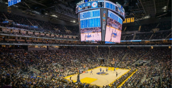 A packed basketball arena with a high-energy atmosphere. The large scoreboard displays game details above the court, surrounded by cheering fans.